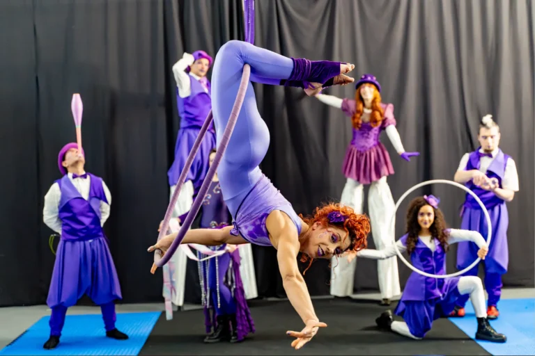 Uśmiechnięta akrobatka artystka z rudymi włosami w fioletowym trykocie wykonuje elastyczny mostek na obręczy aerial hoop. W tle widać grupę akrobatów performerów w fioletowo-purpurowych kostiumach żonglujących maczugami i trzymających koła, scena na macie fitnessowej.