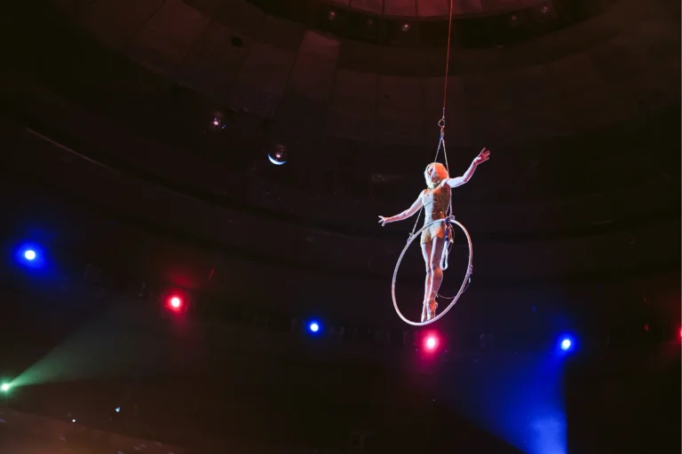 Akrobatka powietrzna w złotym, błyszczącym kostiumie wykonuje widowiskowy numer na kole powietrznym (aerial hoop / lyra). Wisząca wysoko pod kopułą cyrkową, z rozłożonymi ramionami, oświetlona kolorowymi reflektorami.